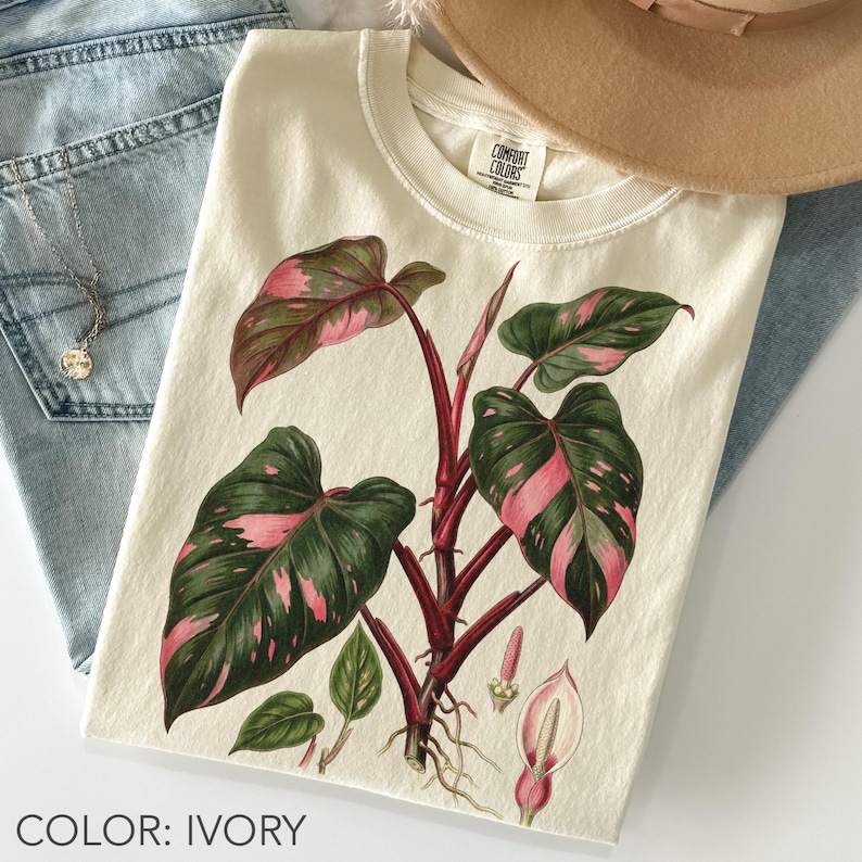 Peut inclure: T-shirt ivoire avec une illustration botanique d'un philodendron princesse rose. Le motif pr&eacute;sente des feuilles vertes avec des panachures roses, des tiges rouges et un dessin botanique d&eacute;taill&eacute;. Le t-shirt est associ&eacute; &agrave; un jean bleu et un chapeau beige.