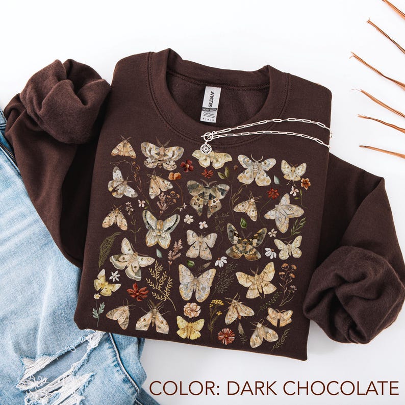May include: A dark chocolate brown crewneck sweatshirt featuring a graphic print of various moths and butterflies in shades of beige, tan, and cream, interspersed with small botanical illustrations of flowers and leaves in muted earth tones.