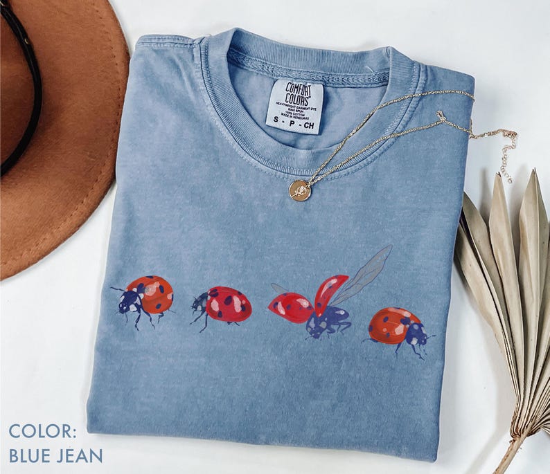 May include: Blue jean Comfort Colors t-shirt with a crew neck and a ladybug graphic. The ladybugs are red with black spots and are in various stages of flight. A gold necklace is draped across the shirt.