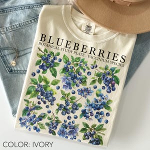 Op de afbeelding: Ivoor kleurig T-shirt met een botanische illustratie van blauwe bessen. Het ontwerp bevat het woord "BLUEBERRIES" in het zwart, met de tekst "BOTANICAL STUDY PLATE. VACCINIUM SPECIES". Het shirt wordt getoond met een blauwe jeans.