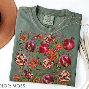 Könnte beinhalten: Ein moosgrünes T-Shirt mit einem Granatapfel-Design, das rote Früchte und orangefarbene Blüten zeigt. Das Shirt hat einen Rundhalsausschnitt und kurze Ärmel. Eine silberne Halskette mit Anhänger ist über dem Shirt drapiert. Der Text "COLOR: MOSS" steht unten.