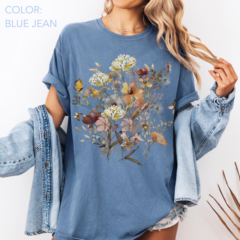 May include: Blue jean colored t-shirt featuring a floral design with butterflies. The graphic includes a variety of flowers in shades of yellow, pink, and brown. The shirt has rolled-up sleeves and a relaxed fit.
