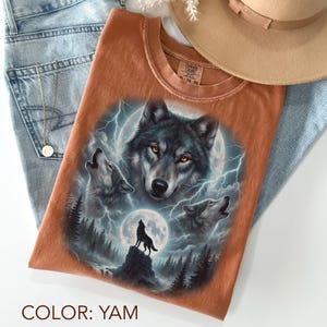 K&ouml;nnte beinhalten: Ein yam-farbenes T-Shirt mit einer detaillierten Grafik von W&ouml;lfen, die den Mond anheulen. Das Design umfasst ein zentrales Wolfsportr&auml;t, drei heulende W&ouml;lfe und die Silhouette eines Wolfs auf einem Felsen. Das Shirt wird mit blauen Jeans und einem beigen Hut kombiniert.