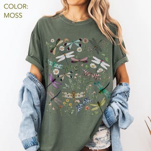 May include: Moss green t-shirt featuring a colorful dragonfly and floral design. The graphic includes various dragonflies in shades of purple, green, blue, and white, along with small flowers and greenery. The shirt has short sleeves and a relaxed fit.