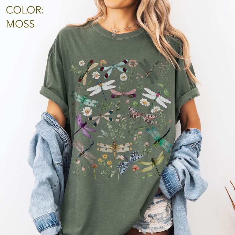 May include: Moss green t-shirt featuring a colorful dragonfly and floral design. The graphic includes various dragonflies in shades of purple, green, blue, and white, along with small flowers and greenery. The shirt has short sleeves and a relaxed fit.