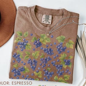 May include: Espresso-colored t-shirt with a crew neck, featuring a pattern of blue grapes and green leaves. The shirt has a Comfort Colors tag and a small silver necklace. The color is labeled as "Espresso".