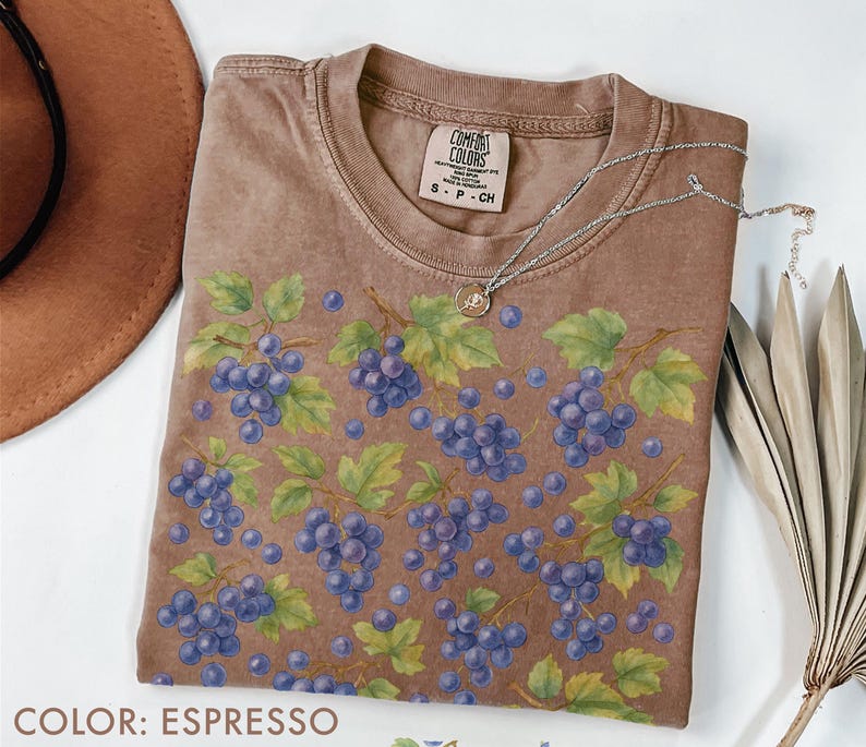 May include: Espresso-colored t-shirt with a crew neck, featuring a pattern of blue grapes and green leaves. The shirt has a Comfort Colors tag and a small silver necklace. The color is labeled as "Espresso".