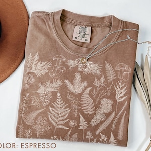 May include: Espresso-colored t-shirt with a crew neck and a botanical design. The shirt features a detailed white illustration of various plants, including ferns, mushrooms, and other foliage. A silver necklace with a pendant rests on the shirt.