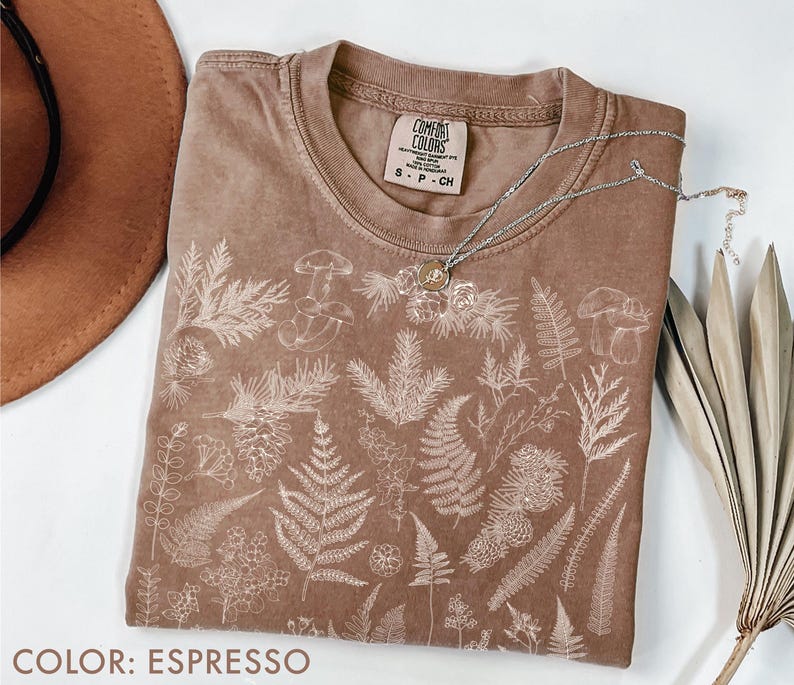 May include: Espresso-colored t-shirt with a crew neck and a botanical design. The shirt features a detailed white illustration of various plants, including ferns, mushrooms, and other foliage. A silver necklace with a pendant rests on the shirt.