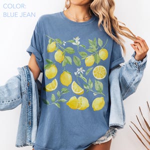 May include: Blue jean colored t-shirt featuring a watercolor design of lemons, leaves, and white flowers. The lemons are whole, sliced, and in various arrangements. The shirt has rolled-up sleeves.