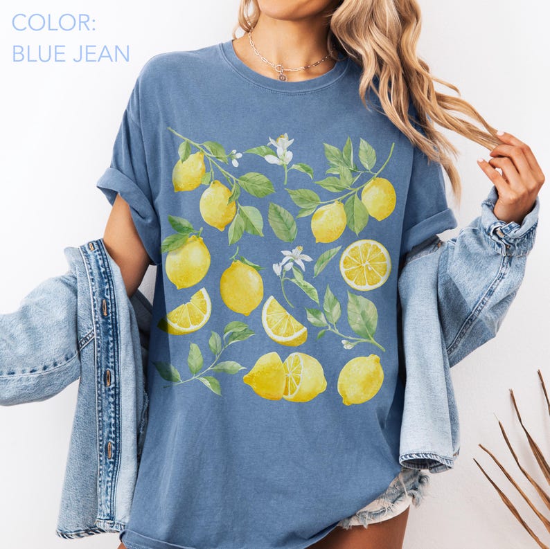 May include: Blue jean colored t-shirt featuring a watercolor design of lemons, leaves, and white flowers. The lemons are whole, sliced, and in various arrangements. The shirt has rolled-up sleeves.