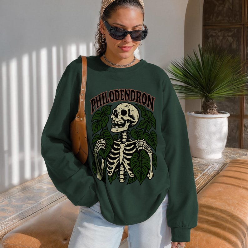 May include: Dark green sweatshirt with the word "PHILODENDRON" above a skeleton graphic surrounded by green leaves. The skeleton is white with black details. The sweatshirt has long sleeves and a crew neck.