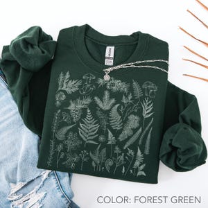 May include: Forest green sweatshirt with a white botanical illustration of ferns, mushrooms, and other plants. A silver necklace with a pendant rests on the sweatshirt. The text "COLOR: FOREST GREEN" is at the bottom of the image.