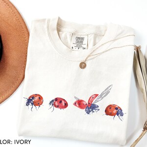 May include: Cream-coloured t-shirt with a watercolour design of four ladybirds in different poses. The ladybirds are red with black spots and blue accents. The shirt has a crew neck and short sleeves. A gold necklace is draped on the shirt.