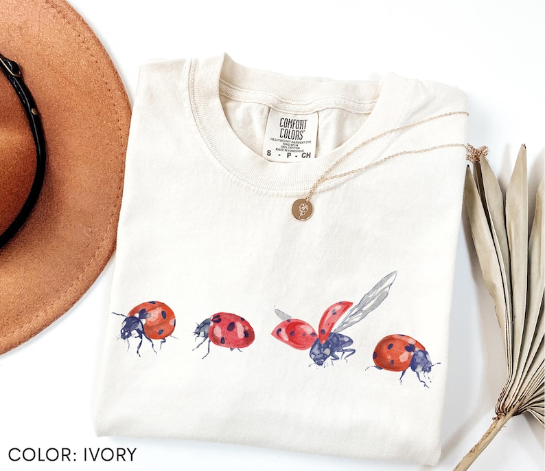 May include: Cream-coloured t-shirt with a watercolour design of four ladybirds in different poses. The ladybirds are red with black spots and blue accents. The shirt has a crew neck and short sleeves. A gold necklace is draped on the shirt.