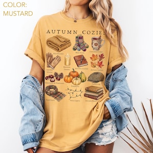 Könnte beinhalten: Ein senfgelbes T-Shirt mit dem Text AUTUMN COZIES und verschiedenen Illustrationen, darunter eine Strickdecke, Wollsocken, ein Becher Apfelwein, Lederhandschuhe, eine Bienenwachskerze, ein Notizbuch, Herbstblätter, Kastanien, Tannenzapfen, Kürbisse, eine Tweed-Mütze, ein karierter Schal, Lichterketten und Bücher.