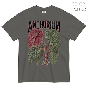 May include: A pepper-coloured t-shirt featuring a graphic print of an Anthurium plant with large, textured green and reddish-maroon leaves, visible roots, and the word ANTHURIUM printed in a bold, dark, distressed font above the botanical illustration.