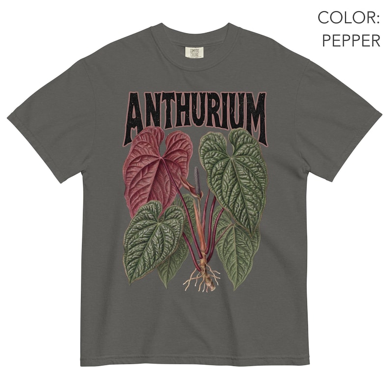 May include: A pepper-coloured t-shirt featuring a graphic print of an Anthurium plant with large, textured green and reddish-maroon leaves, visible roots, and the word ANTHURIUM printed in a bold, dark, distressed font above the botanical illustration.