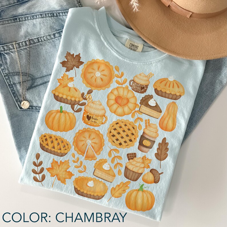 May include: A light blue chambray t-shirt with a fall-themed design. The graphic features pumpkins, pies, leaves, and coffee cups. The shirt is folded on blue jeans, with a gold necklace and a brown hat in the background. The shirt's colour is described as chambray.