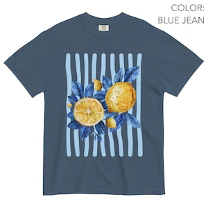 May include: Blue jean colored t-shirt featuring a watercolor design of lemons and blue leaves. The design includes a sliced lemon, a whole lemon, and light blue vertical stripes. The shirt has a classic crew neck and short sleeves.