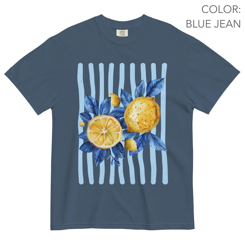 May include: Blue jean colored t-shirt featuring a watercolor design of lemons and blue leaves. The design includes a sliced lemon, a whole lemon, and light blue vertical stripes. The shirt has a classic crew neck and short sleeves.