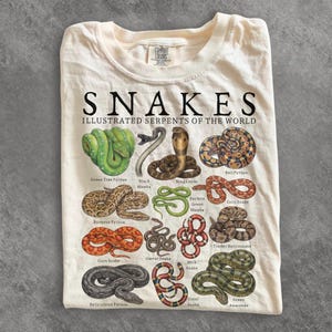 Könnte beinhalten: Ein cremefarbenes T-Shirt mit dem Text SNAKES ILLUSTRATED SERPENTS OF THE WORLD und detaillierten Illustrationen verschiedener Schlangenarten, darunter ein Grüner Baumpython, eine Königskobra und ein Königspython, mit ihren Namen darunter.