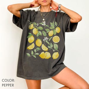 May include: Pepper-colored t-shirt featuring a watercolor design of lemons, leaves, and blossoms. The design includes whole and sliced lemons, with green leaves and white blossoms. The shirt is a relaxed fit.