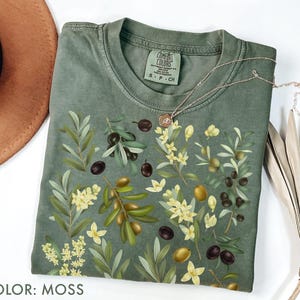 May include: Moss green t-shirt with an olive branch design. The shirt has a crew neck and short sleeves. The olive branch design includes green leaves, black and yellow olives, and small white flowers. A gold necklace is draped over the shirt.