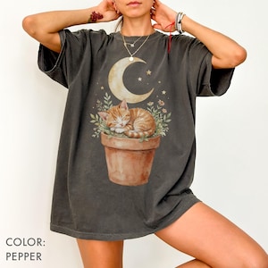 May include: Pepper-colored t-shirt featuring a watercolor illustration of a sleeping orange tabby cat in a terracotta pot, with a crescent moon and stars in the background. The shirt's color is labeled as "Pepper".