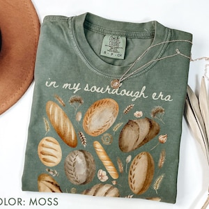 May include: Moss green t-shirt with the phrase "in my sourdough era" and illustrations of various breads. The shirt has a crew neck and short sleeves. A silver necklace with a small pendant is draped across the shirt.