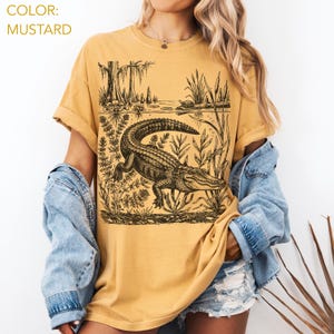 May include: Mustard-colored t-shirt featuring a detailed black and white illustration of an alligator in a swamp setting. The design includes aquatic plants, trees, and the text "COLOR: MUSTARD". The shirt has short sleeves and a relaxed fit.