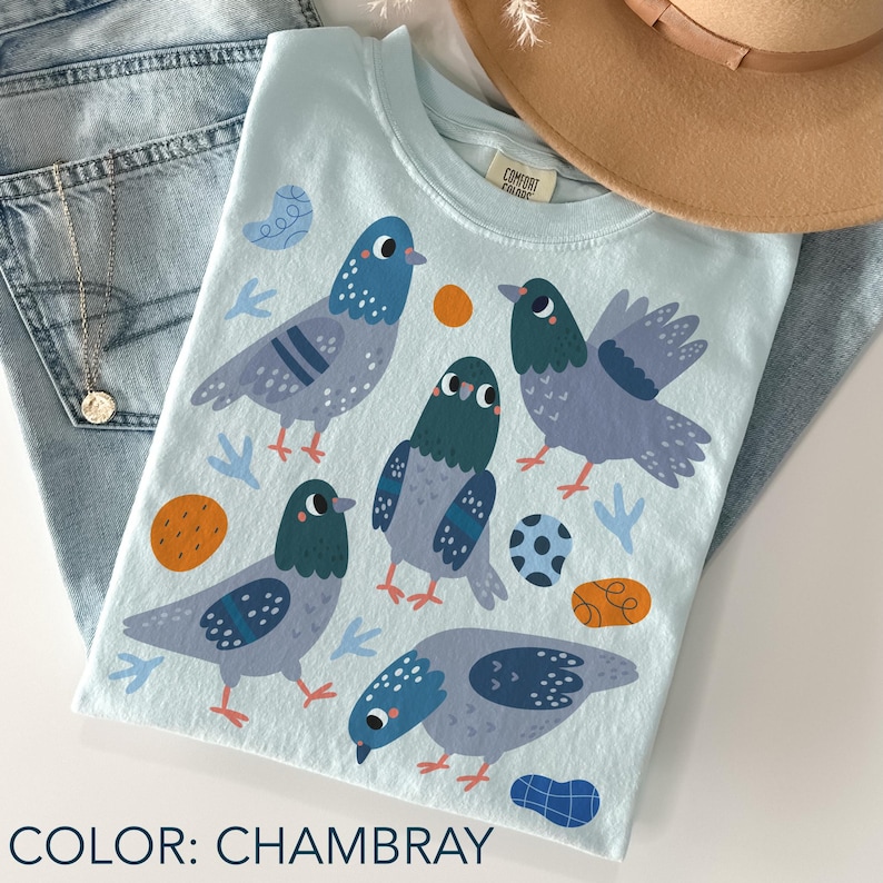 May include: A light blue t-shirt with a graphic of five cartoon pigeons in shades of gray and blue, with orange and blue accent shapes. The shirt is labeled "COLOR: CHAMBRAY".