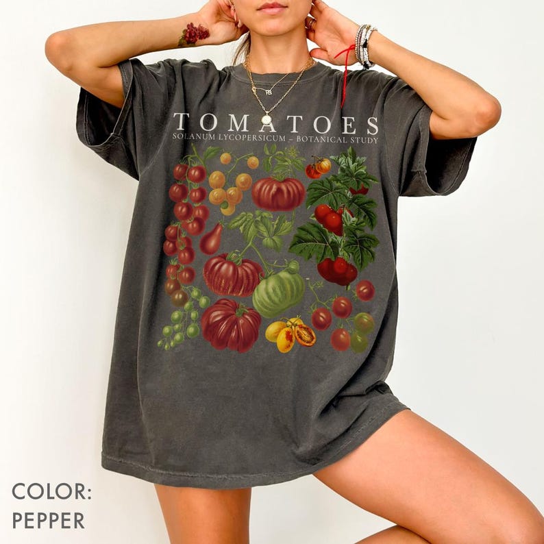 May include: Dark grey t-shirt featuring a botanical illustration of various tomatoes. The shirt displays the word "TOMATOES" above the illustration, with "SOLANUM LYCOPERSICUM - BOTANICAL STUDY" below. The shirt is a relaxed fit.