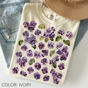 May include: Ivory-colored t-shirt featuring a pattern of purple pansies with green leaves. The shirt is a Comfort Colors brand. The text "COLOR: IVORY" is at the bottom of the image. A necklace, jeans, and a hat are also visible.