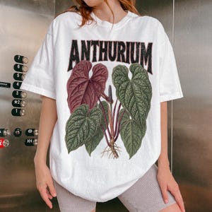 May include: A white short-sleeve t-shirt featuring a graphic print of an Anthurium plant with large green and deep red heart-shaped leaves. The word ANTHURIUM is printed in a bold, distressed black font above the botanical illustration.