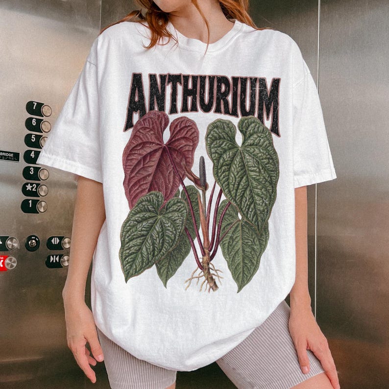 May include: A white short-sleeve t-shirt featuring a graphic print of an Anthurium plant with large green and deep red heart-shaped leaves. The word ANTHURIUM is printed in a bold, distressed black font above the botanical illustration.