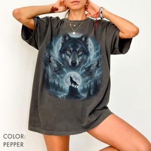K&ouml;nnte beinhalten: Pfefferfarbenes T-Shirt mit einem Wolf-Motiv. Das Design zeigt ein zentrales Wolfsgesicht, drei heulende W&ouml;lfe, einen Vollmond und eine Waldszene mit Blitzen. Das T-Shirt hat kurze &Auml;rmel und eine lockere Passform.