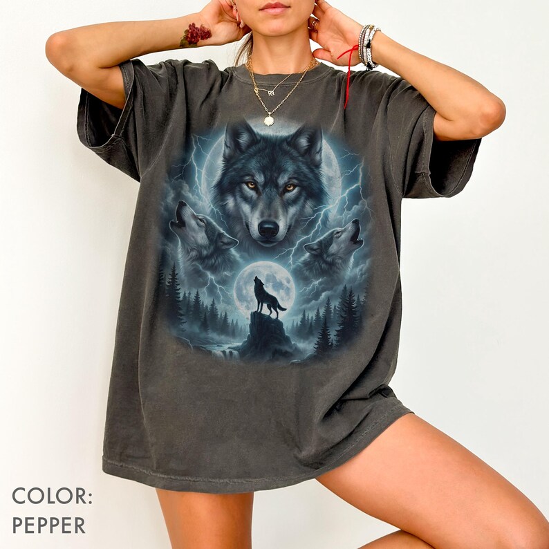 K&ouml;nnte beinhalten: Pfefferfarbenes T-Shirt mit einem Wolf-Motiv. Das Design zeigt ein zentrales Wolfsgesicht, drei heulende W&ouml;lfe, einen Vollmond und eine Waldszene mit Blitzen. Das T-Shirt hat kurze &Auml;rmel und eine lockere Passform.