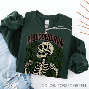May include: Forest green sweatshirt with a skeleton graphic surrounded by leaves and the word "PHILODENDRON" in brown lettering. A silver necklace with a circular pendant rests on the sweatshirt. The sweatshirt is folded on top of a pair of blue jeans.
