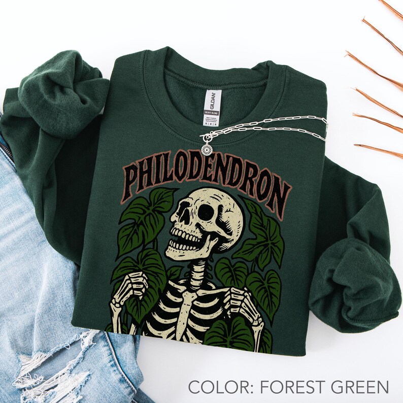 May include: Forest green sweatshirt with a skeleton graphic surrounded by leaves and the word "PHILODENDRON" in brown lettering. A silver necklace with a circular pendant rests on the sweatshirt. The sweatshirt is folded on top of a pair of blue jeans.