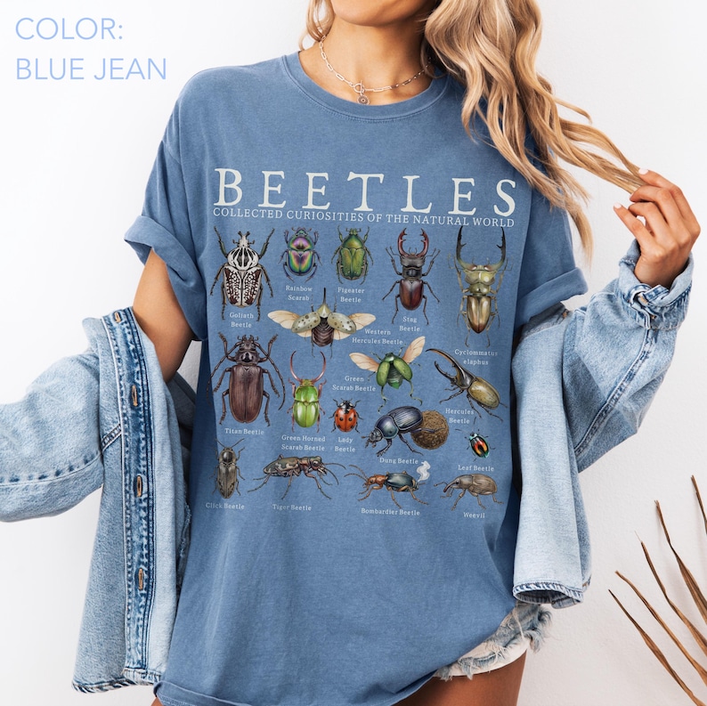 Puede incluir: Camiseta azul vaquero con la palabra "BEETLES" y el texto "COLLECTED CURIOSITIES OF THE NATURAL WORLD". La camiseta presenta ilustraciones de varios escarabajos. Tiene mangas cortas y un corte relajado.