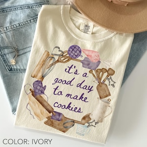 May include: Ivory t-shirt featuring a watercolour design of baking tools and the phrase "It's a good day to make cookies" in purple script. The shirt is shown with blue jeans and a necklace.