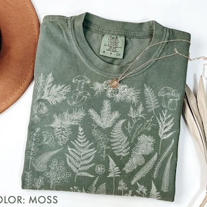 May include: A moss-colored t-shirt with a botanical design featuring ferns, mushrooms, and other plants. The shirt has a crew neck and short sleeves. A silver necklace with a circular pendant rests on the shirt. The text "COMFORT COLORS" is visible on the tag.