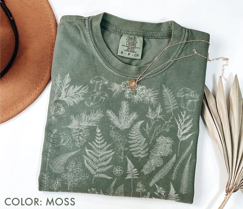 May include: A moss-colored t-shirt with a botanical design featuring ferns, mushrooms, and other plants. The shirt has a crew neck and short sleeves. A silver necklace with a circular pendant rests on the shirt. The text "COMFORT COLORS" is visible on the tag.
