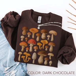 Puede incluir: Sudadera de color chocolate oscuro con ilustraciones de varios hongos en tonos marrón, naranja y amarillo. La sudadera tiene cuello redondo y mangas largas. Un collar plateado con un colgante circular está drapeado en la parte delantera. El texto "COLOR: DARK CHOCOLATE" está en la parte inferior.