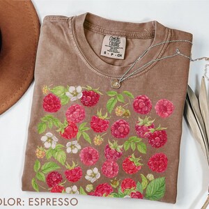 Op de afbeelding: Espresso-kleurig T-shirt met een frambozenontwerp. De afbeelding toont gedetailleerde illustraties van rode frambozen, witte bloemen en groene bladeren. Het shirt heeft een ronde hals en korte mouwen. Een zilveren ketting met een kleine hanger ligt op het shirt.