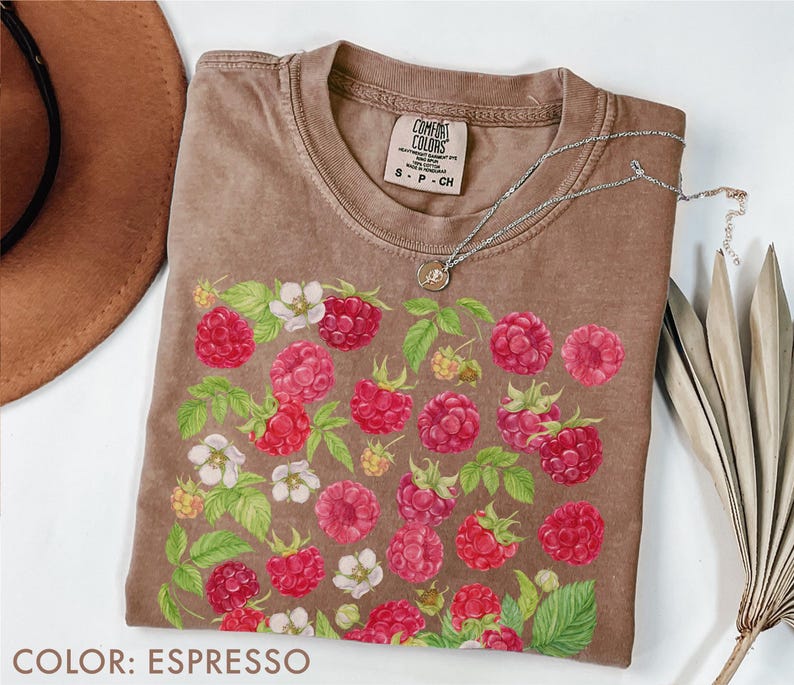 Op de afbeelding: Espresso-kleurig T-shirt met een frambozenontwerp. De afbeelding toont gedetailleerde illustraties van rode frambozen, witte bloemen en groene bladeren. Het shirt heeft een ronde hals en korte mouwen. Een zilveren ketting met een kleine hanger ligt op het shirt.