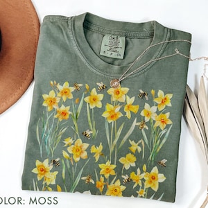 Puede incluir: Camiseta verde musgo con un diseño de acuarela de narcisos amarillos y abejas. La camiseta tiene cuello redondo y mangas cortas. Un delicado collar plateado con un pequeño colgante descansa sobre la camiseta. El color de la camiseta está etiquetado como "MOSS".
