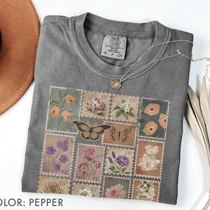K&ouml;nnte beinhalten: Pfefferfarbenes T-Shirt mit Rundhalsausschnitt und floralem Stempeldesign. Das Design umfasst Bilder von Blumen und Schmetterlingen. Das Shirt hat ein Etikett mit der Aufschrift "COMFORT COLORS" und "S P CH". Eine silberne Halskette mit Anh&auml;nger ist auf dem Shirt drapiert.