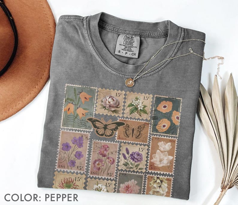 K&ouml;nnte beinhalten: Pfefferfarbenes T-Shirt mit Rundhalsausschnitt und floralem Stempeldesign. Das Design umfasst Bilder von Blumen und Schmetterlingen. Das Shirt hat ein Etikett mit der Aufschrift "COMFORT COLORS" und "S P CH". Eine silberne Halskette mit Anh&auml;nger ist auf dem Shirt drapiert.